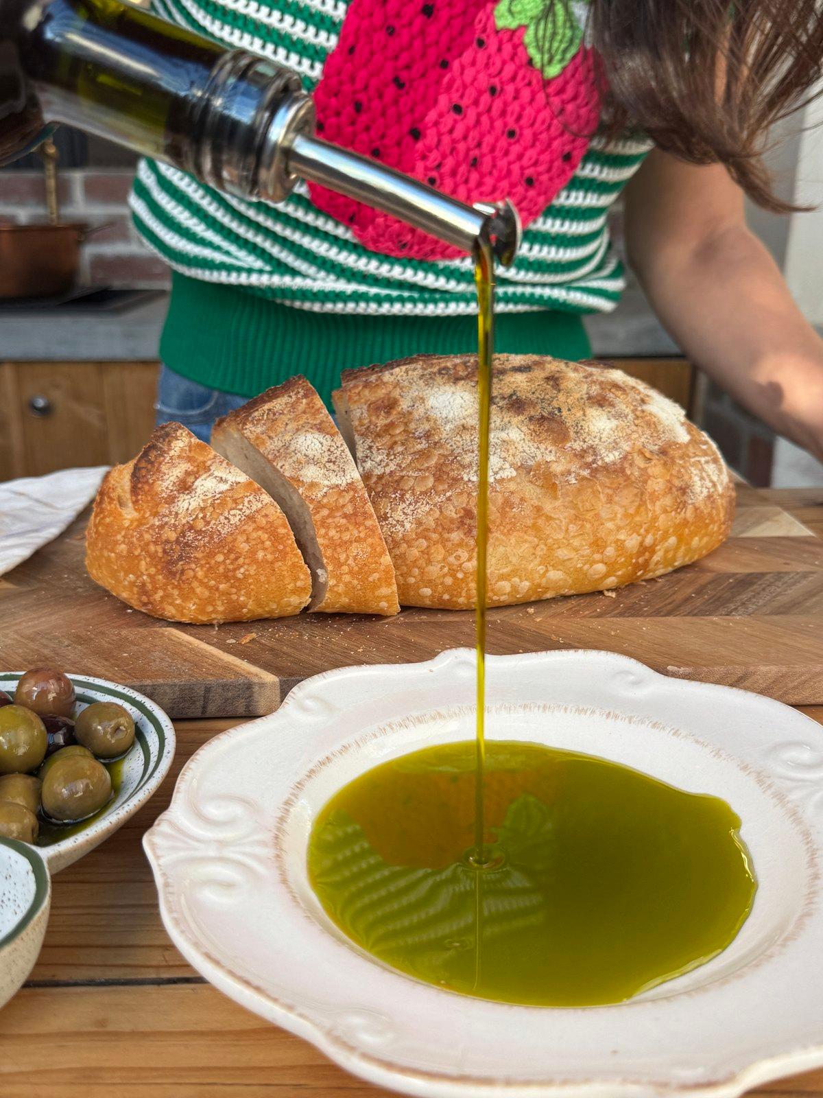 Olive oil poured into a dish beside a sliced sourdough loaf and olives